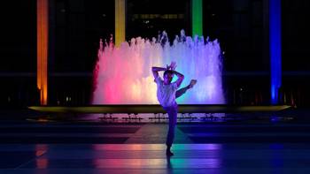 A dancer poses with a dancing figure in front of a fountain which is illuminated by four columns of light, each with a different colour